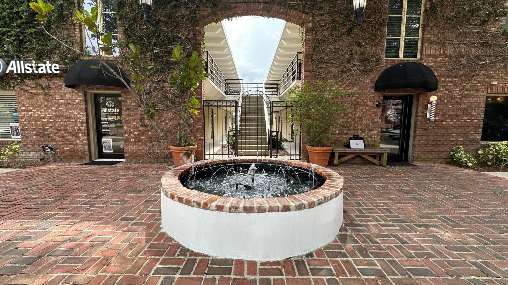 Circular brick fountain in a courtyard with a two-story staircase behind a gate; surrounded by a red brick building with ivy, an Allstate office on the left, a barber shop on the right, and a bench with a laptop bag.
